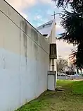 Vue de côté d'un bâtiment en béton, avec des traces d'humidité ; le bâtiment se prolonge avec une partie montante surplombée d'une croix.