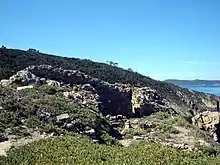 Photographie couleur de ruines dans la garigue. La mer semble surplomblée.