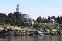Sur une berge partiellement boisée, une maison de 2 étages en bois, d'où dépasse le court cylindre du phare.