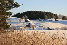 Photographie au premier plan d'un lac gelé avec un esker le bordant sur lequel se trouvent quelques maisons en bois, le tout dans un passage hivernal.