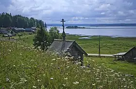 Vue depuis une petite colline d'une petite chapelle en bois en premier plan d'architecture russe avec une croix orthodoxe à son sommet. En second plan à gauche se distingue un village avec des maisons en bois, et en arrière-plan le lac avec au fond des collines recouvertes de taïga.