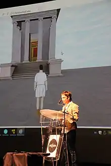 Une jeune femme parlant à un pupitre, avec à l'arrière-plan un personnage personnage virtuel donnant l'échelle des édifices restitués.