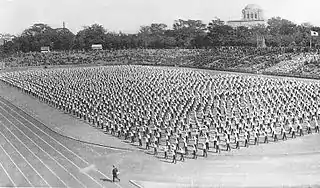 Photo noir et blanc de rangeés de personnes accomplissant, à l'unisson, des exercices physiques, au centre d'un stade.