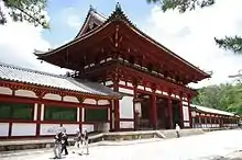 Porte dans le mur d’enceinte, aux poutres rouges et aux murs blancs. Toit à demi-croupes au faîte plat, courbé à l’extrémité.