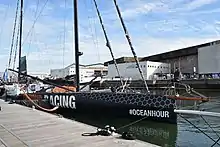 Voilier noir amarré au ponton. On voit le foil tribord.