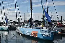 bateau bleu et blanc vu de 3/4 avant, amarré au ponton. En arrière plan, on voit l'avant d'un autre Imoca, Corum L'Épargne.