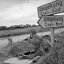 Photo en noir et blanc de deux soldats à couvert, en position dans un fossé près d'une route, sous un panneau routier écrit en allemand et indiquant la direction de Caen.