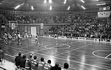 Photo en noir et blanc d'un match de basket. On distingue les remplaçants de Denain assis sur le banc au bord du terrain regardant les joueurs des deux équipes sous le panier.