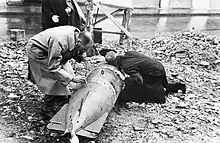 Deux hommes en civil penchés sur une bombe, parmi des gravats. L'un d'eux a l'oreille posée dessus, l'autre utilise un stéthoscope.