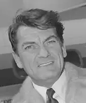 Photo en noir et blanc d'un élégant quinquagénaire souriant à l'objectif. Un avion à l'arrière-plan.