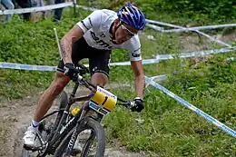 Homme descendant une pente à VTT, portant un casque bleu, un maillot blanc et un cuissard noir.