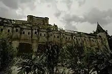Photo couleur d'un rempart en brique rouge à trainées vertes de mousse. Le mur taluté en bas comporte des contreforts en demi-cylindre et le haut est doté d'une rangée de fenêtres sous le chemin de ronde. La tour à droite est polygonale surmontée d'un toit pointu, également en brique.