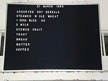 Tableau d’affichage Lectovisuel noir rainuré, en matière synthétique, encadré d’aluminium, sur lequel on a accroché des lettres (certaines manquent) pour donner le menu du 21 mars 1963 : assortiment de céréales sèches, blé entier à la vapeur, 1 œuf brouillé, 2 lait, compote de fruits, pain grillé, pain, beurre, café.