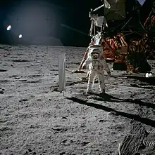 Photo d'un astronaute sur la Lune devant un LEM avec sur sa gauche un collecteur de vent solaire.