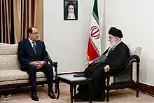 Deux homme assis en discussion, l'un en costume, l'autre en barbe blanche et turban