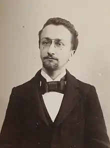 Photographie en noir et blanc d'un homme en costume complet portant une courte barbe et des lunettes.
