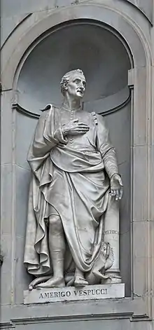 Photographie d'une statue en pied d'un homme assez âgé la main droite sur la poitrine. À ses pieds se trouve une sorte d'iguane.