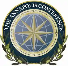 Un logo circulaire, semblable à un sceau. Au centre une rose des vents jaune superposée à un globe stylisé bleu. Autour, une couronne jaune avec les quatre points cardinaux. Autour, une bande bleue cerclée de jaune avec écrit The Annapolis Conference. En bas, des rameaux d’olivier épousent la forme du sceau.