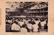 Photo en noir et blanc montrant une foule assise, hommes habillés en blanc, devant un grand bâtiment à un étage (le palais royal). Au fond à gauche se tiennent des marins français et un groupe d'officiers. Enfin, contre le mur du palais, les dignitaires wallisiens (Lavelua, chefs), l'évêque et le résident de France.
