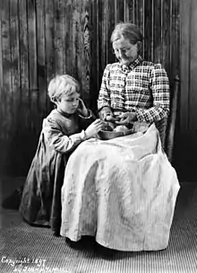 Portrait en noir et blanc d’une femme âgée et d’un enfant posant devant un lambris de bois : la femme, assise, épluche au couteau une pomme de terre au-dessus d’un plat posé sur ses genoux tandis que l’enfant, à genoux sur sa droite, examine un tubercule du récipient. Le maintien de la femme est guindé mais le visage incliné et attentif est légèrement souriant ; elle porte des lunettes rondes et les cheveux gris tirés en chignon, un corsage à carreaux et un ample tablier clair, ligné, qui descend jusqu’au sol. L’enfant aux cheveux courts et blonds, semble très sérieux ; il porte un long tablier à carreaux vichy dont les longues manches sont resserrées aux poignets.