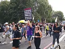 Une foule d'hommes et de femmes marchent dans une rue urbaine, avec des arbres en arrière-plan. Certaines personnes portent des pancartes