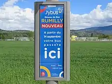 Photographie en couleurs d’un panneau gris et bleu au milieu des champs verts, qui marque un arrêt de bus