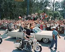 Une limousine blanche avec trois personnes à l'arrière et deux à l'avant. Deux policiers à l'avant-plan et une foule à l'arrière-plan.