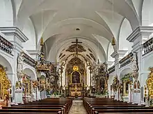 Une haute salle voûtée aux murs blancs et à la riche décoration dorée, avec des rangées de bancs de chaque côté d'une allée centrale.