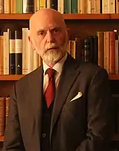 photo d'un barbu vétu d'un costume trois-pièces posant devant une bibliothèque.