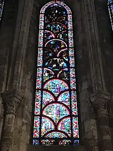 Vitrail composé de pièces arrondies en forme de coquillages. Sont représentés différents personnages dans plusieurs scénettes dont, en bas, un homme habillé de rouge s'agenouillant devant deux hommes levant le bras et semblant le menacer.  Les parties hautes sont difficilement visibles en raison du noircissement du vitrail.