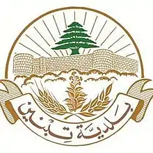 Description de Blason de Tibnine : Sur un fond blanc et en couleur vieux or, la partie supérieure des rayons de soleil en demi-cercle jaillissent de derrière le Toron des Chevaliers stylisé surplombé par un Cèdre vert du Liban. A la hauteur des fondations du château fort les sommets des arbres séparent le blason en deux. Une banderole ferme la partie inferieure portant l’inscription couleur bronze en arabe, municipalité de Tibnine. Dans son arrondi à dextre couché vers la droite, un rameau d'olivier en fruits, au centre une pousse de tabac à maturité, à sénestre couché vers la gauche, un épi du blé.