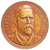 Médaille représentant un homme barbu, aux cheveux courts et portant des lunettes. Sur la gauche l'inscription OLIVER C. et sur la droite BOSBYSHELL.
