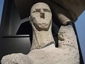 Une statue partiellement reconstituée en pierre crème, accrochée à un portoir noir, le bouclier au-dessus de la tête.