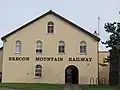 La façade d'un bâtiment aux murs ocre avec six fenêtres en ogive. Les mots « BRECON MOUNTAIN RAILWAY » y sont écrits en grandes lettres marron.