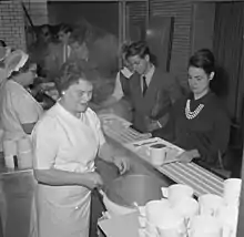 Photographie en noir et blanc. Des femmes en uniforme blanc servent les portions de nourriture sur les plateaux qu’étudiants et étudiantes font glisser devant elles sur un comptoir rainuré. Les étudiants sont en costume-cravate, les étudiantes sont élégamment vêtues.