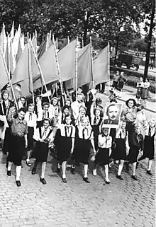 Photo noir et blanc d'un groupe de jeunes filles en chemise blanche et juppe noire, défilant dans une rue tout en agitant des drapeaux et le portrait d'un homme.