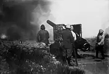 Photographie d'un canon lourd tirant depuis une position légèrement protégée par des sacs de sable. Plusieurs artilleurs se tiennent à proximité.