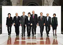 Photographie d'un groupe de cinq hommes et trois femmes marchant vers la caméra sur un fond blanc et sur un sol représentant le drapeau suisse.