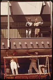 Photographie des années 1970 montrant deux ouvriers travaillant en sous-face de passerelle tandis que deux personnes se tiennent à l'intérieur de la passerelle vitrée.
