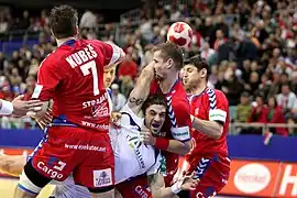 Bertrand Gilles, pratiquement à l'horizontale, tenu par 3 adversaires. Il tente de tirer avec le bras droit en l'air.