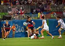 Au centre, de face, une athlète avec le ballon sous le bras droit. Avec le bras gauche, elle s'apprête à repousser une adversaire.