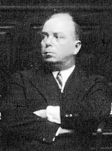 Photographie en noir et blanc et en buste d'un homme entre deux âges en costume sombre, les bras croisés et regardant sur sa droite