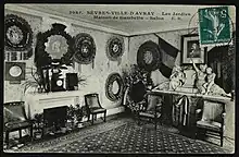 Carte postale en noir et blanc oblitérée d'un timbre vert montrant l'intérieur d'un salon comprenant des tableaux et des portraits photographiques accrochés au mur, ainsi que des objets d'art disposés sur des meubles et une petite cheminée.