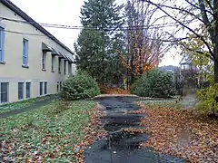 Une allée dans un jardin longeant un bâtiment. L'allée est partiellement recouverte de feuilles mortes.