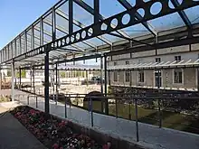 Photographie présentant un déambulatoire quadrangulaire dallé recouvert d'une structure métallique légère supportant une verrière.