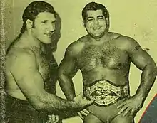 Photo en noir et blanc. Un catcheur est à gauche, Bruno Sammartino. Il porte un slip de catch foncé, est torse nu et à des cheveux mi-longs noirs. Il pointe le titre du catcheur de droite en regardant et en semblant parler à quelqu'un à droite. Le catcheur de droite, Pedro Morales, porte un slip clair, la ceinture de champion et à des cheveux mi-longs noirs. Il a les mains sur ses hanches et rit.