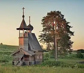 Vue au couchant d'une chapelle en bois qui possède un petit beffroi et un toit en tente, le tout d'une architecture en bois russe.