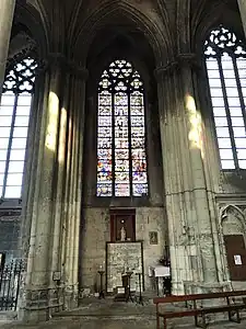 Grand vitrail avec des teintes dominantes froides (bleu et vert surtout). Cinq personnages sont présents. Au centre de l'image, une femme avec une auréole porte une croix et lance des roses. En bas à gauche de l'image, un poilu de la Grande surmonte un marin en train de prier. À leur droite, un homme barbu tonsuré surmonte une femme à la peau mate en train de prier. Tout en bas, des écritures sont entourées de deux blasons.