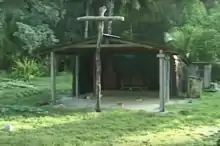 Photo d'un bâtiment en bois, sans mur sur le devant avec à l'entrée une grande croix en bois, à l'arrière-plan la forêt.