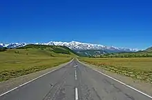 Photographie depuis le centre de la route. Le paysage est verdoyant, avec en arrière-plan des cimes enneigées, dans la steppe de Kouraï.
